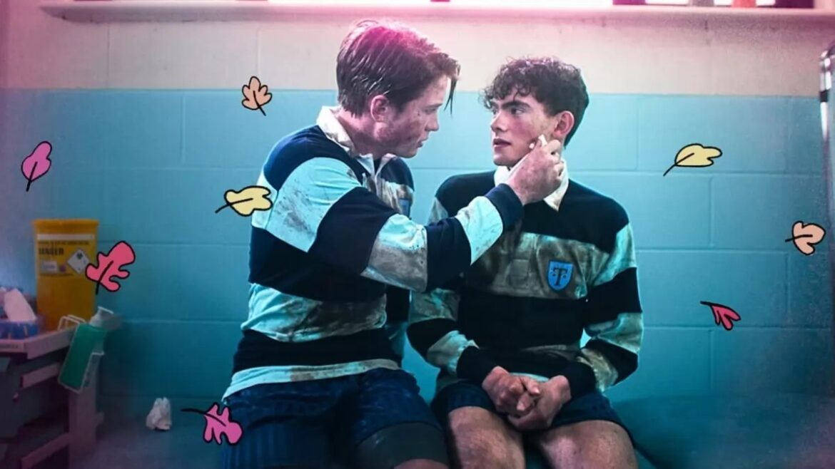 IMAGE 1691095929 16×9 Nick Nelson played by Kit Connor gently wipes the cheek of Charlie Spring played by Joe Locke. Both young men are wearing rugby uniforms. There's a gentle pink aura and cartoon oaks leaves over the picture.
