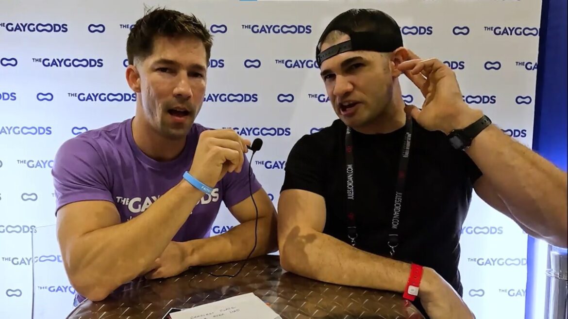 Kyler Drayke, a caucasian man with a black baseball cap worn backwards is gesturing to his left ear with his left hand while describing kissing the ears. Eddie Patrick, to his right, has his mouth agape listening to Kyler while facing the camera. Eddie is wearing a purple T-shirt saying 'The Gay Goods' in white lettering. The men are speaking in front of a white background with 'The Gay Goods' written with purple lettering.