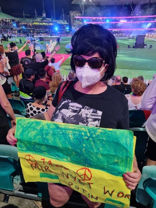 Barbara Karpinski is holding a green and yellow poster. She is an older woman with black hair in a small beehive, dark glasses and wearing a white N95 respirator. In the background people are seen waving at the celebrations on the cricket pitch.