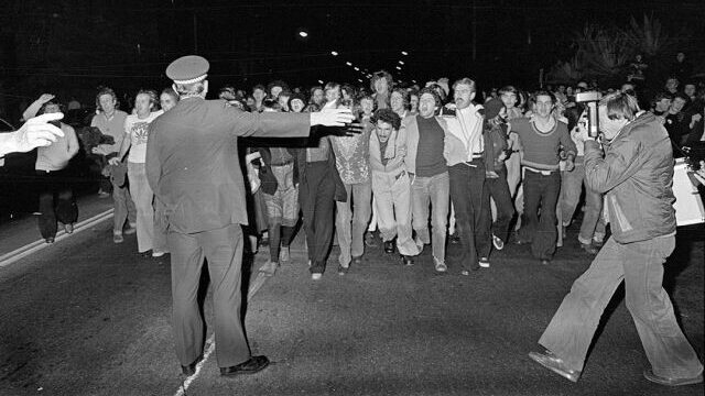 A horde of gay liberation marchers heading towards police officers facing away from the camera pointing to the right. The photograph is in black and white.