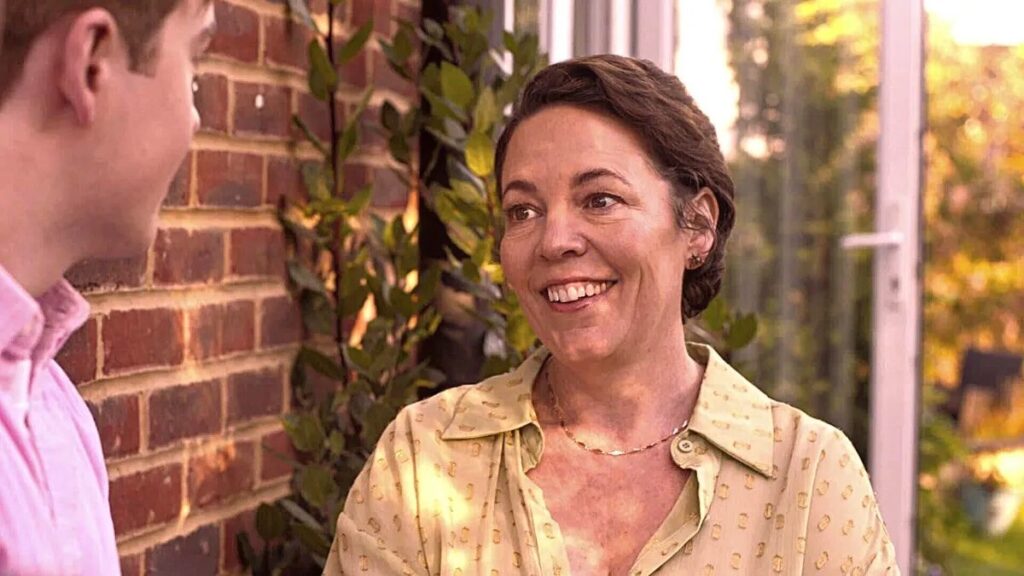 Sarah Nelson (Olivia Colman) is smiling to an off-camera Nick Nelson (Kit Harrington). She is wearing a yellow top and she is standing in a conversatory with the late afternoon light dancing over her face.