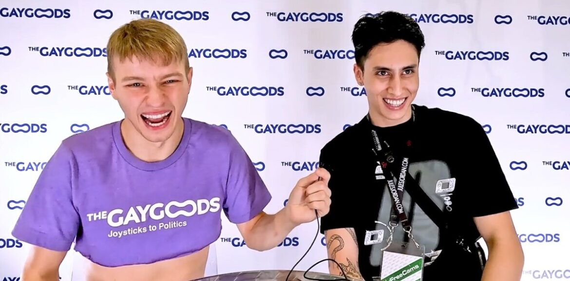 Noah, a blonde twink with a purple crop top saying 'The Gay Goods: Joysticks to Politics' is laughing facing the camera speaking with Angel Elias. Angel Elias is a Latinx young man smiling wickedly at an off-camera Alex Myers (unpicture) in a black T-shirt. Noah is holding a small lapel mic in his hand pointing it in Angel's direction.