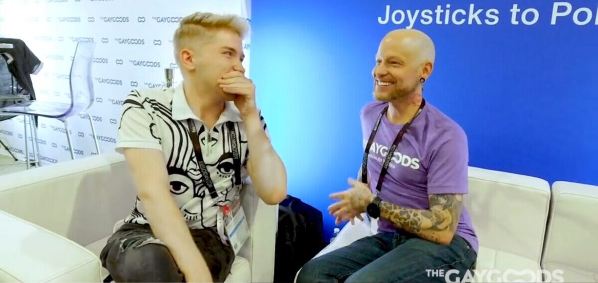 Screenshot 2024 03 07 131408 Zacc Andrews a blonde young males is giggling with his hand over his mouth speaking with Justin (aka Disco Dollie) a tattooed man with a purple T-shirt saying 'The Gay Goods'