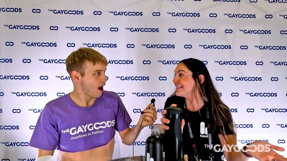 Noah White,. a blonde twink ina purple crop top has his mouht open in shock next to maria Luna, a young brunette woman as she explains 'The Handy' a black automated stroking machine. Behind them is a white banner with the words The Gay Goods repeated in blue lettering.