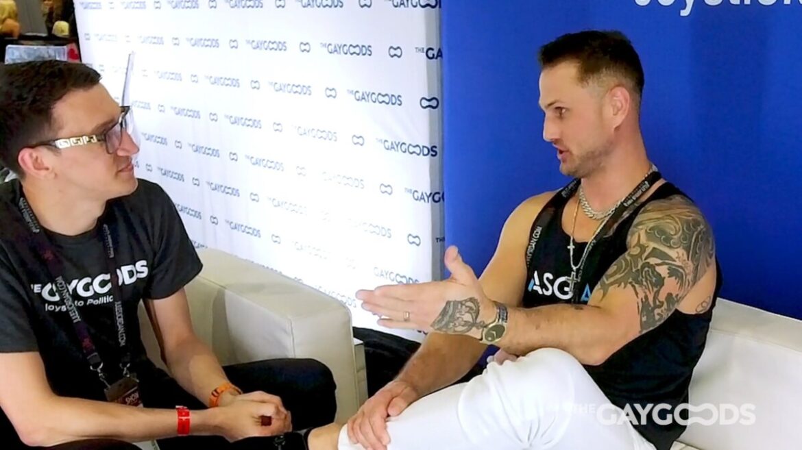 Screenshot 2024 03 15 222743 Blain O'Connor, a tattooed muscular man with a black tank top (with ASG Max written in white and blue lettering on it) speaks with Alex Sparks, a thin man with glasses, black hair and a blakc t-shirt with The Gay Goods written with white lettering. They are sitting on white leather couches.