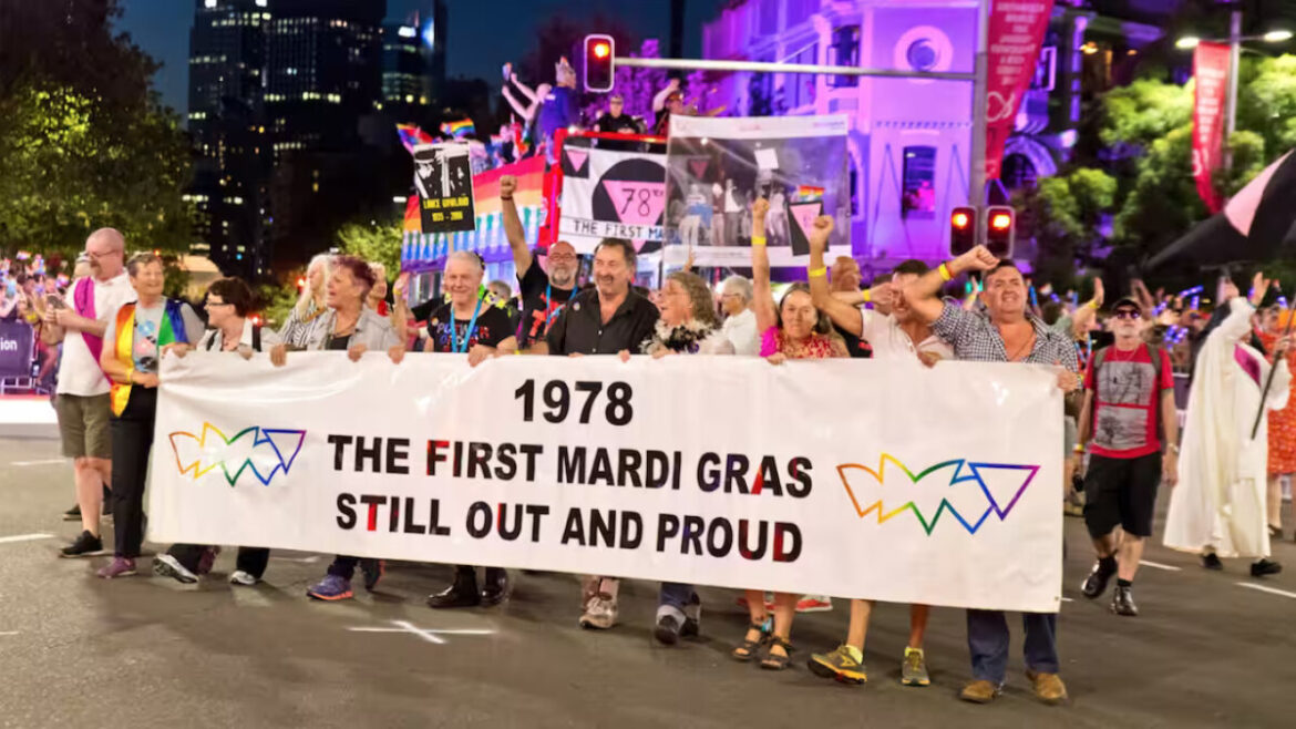 A contingent of older people with holding a banner saying "1978 The First Mardi Gras Still Out And Proud" with rainbow outlines of the Sydney opera house and an upside-down triangle. They are marching in the Sydney Mardi Gras Parade. Many have their right fists in the air in a sign of solidarity and defiance. In the background a banner with a pink upside down triangle says "78ers" on a double decker float with people waving rainbow flags.