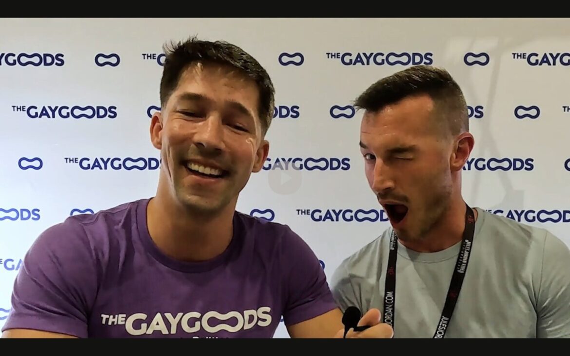 Eddie Patrick, a caucasian male with dark hair is smiling widely. He is wearing a purple shirt with the words 'The Gay Goods' written onto it in white. Derek Kage is sitting to his left. Derek is a late-30s with short cropped dark hair and a moustache. He is winking histrionically at the viewer.