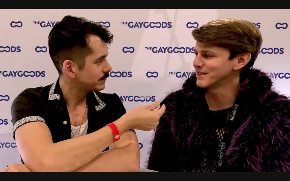 Jonah Wheeler, a moustached man with dark brown hair is holding a lapel mic to Sage Roux. Sage Roux has blonde hair and is wearing a deep purple faux fur. Jonah is wearing a short sleeved black shirt with a patterned lining over a white singlet and silver chain. He is staring at Sage, who is smiling gently at him is response.