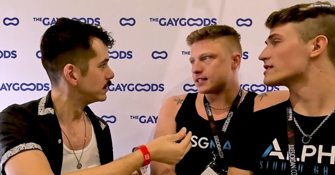 Screenshot 2024 05 10 142538 Jonah Wheeler is speaking with Logan Aarons and Kam Stone. Jonah Wheeler is a moustached man with a black short sleeved shirt, holding a lapel microphone in his right hand. Logan Aarons is a blonde man with a black tank top his right arm on the shoulder of his partner Kam Stone. Kam has short hair and is wearing a black t-shirt.