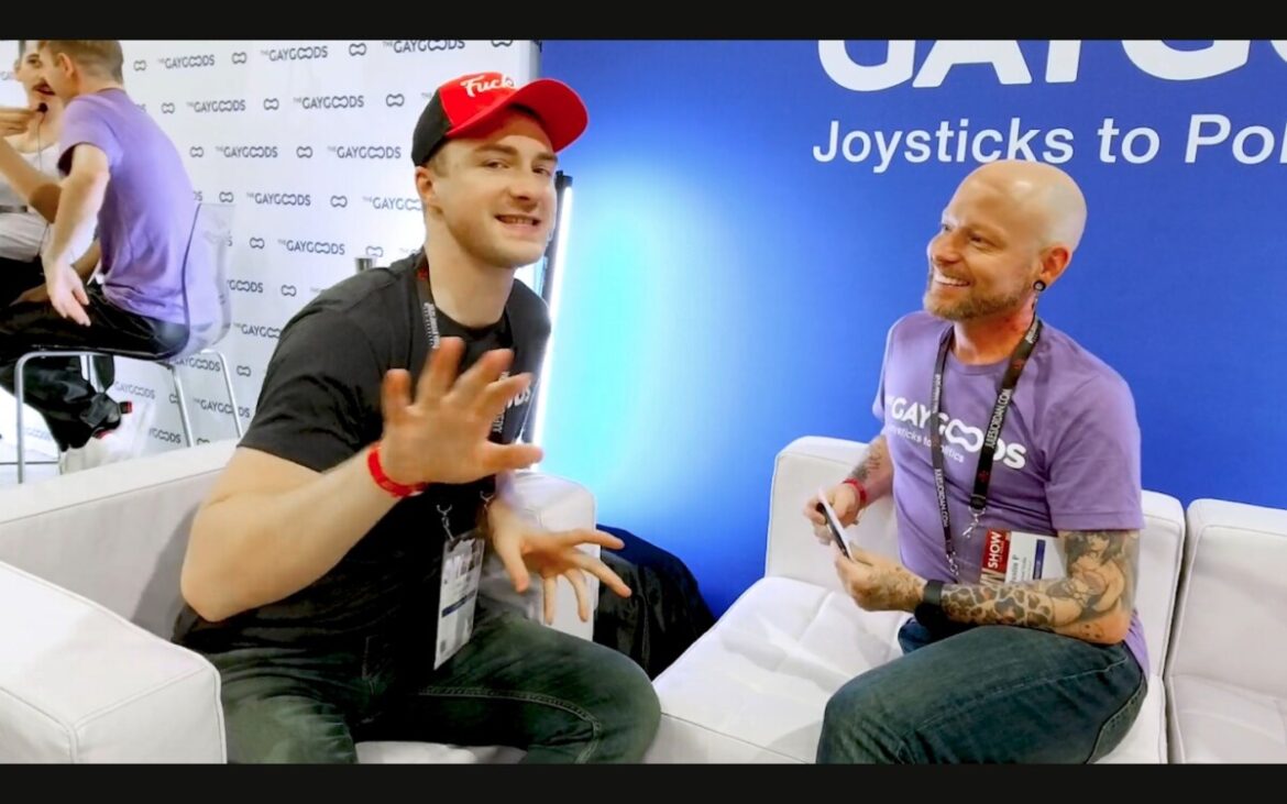 Cole Blue is a caucasian man wearing a black t-shirt and red cap saying "Fuck Boy". He is gesturing with his hand to the viewer. He is sitting on a white leather chair across from Justin Carnal, a tattooed bald man with a wide bright smile and purple t-shirt. They are sitting in front of a blue background.