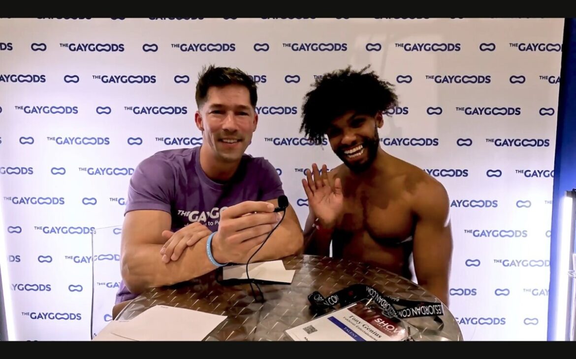 Eddie Patrick (a caucasian man with black short cropped hair and a purple T-shirt) holds a lapel microphone in his right hand. To his left is Tony Genius (an African-American man, shirtless with a bouncy afro) is smiling and waving. He is waving with his right hand. They are sitting in front of a backdrop with 'The Gay Goods' written with blue letters on a white backdrop.