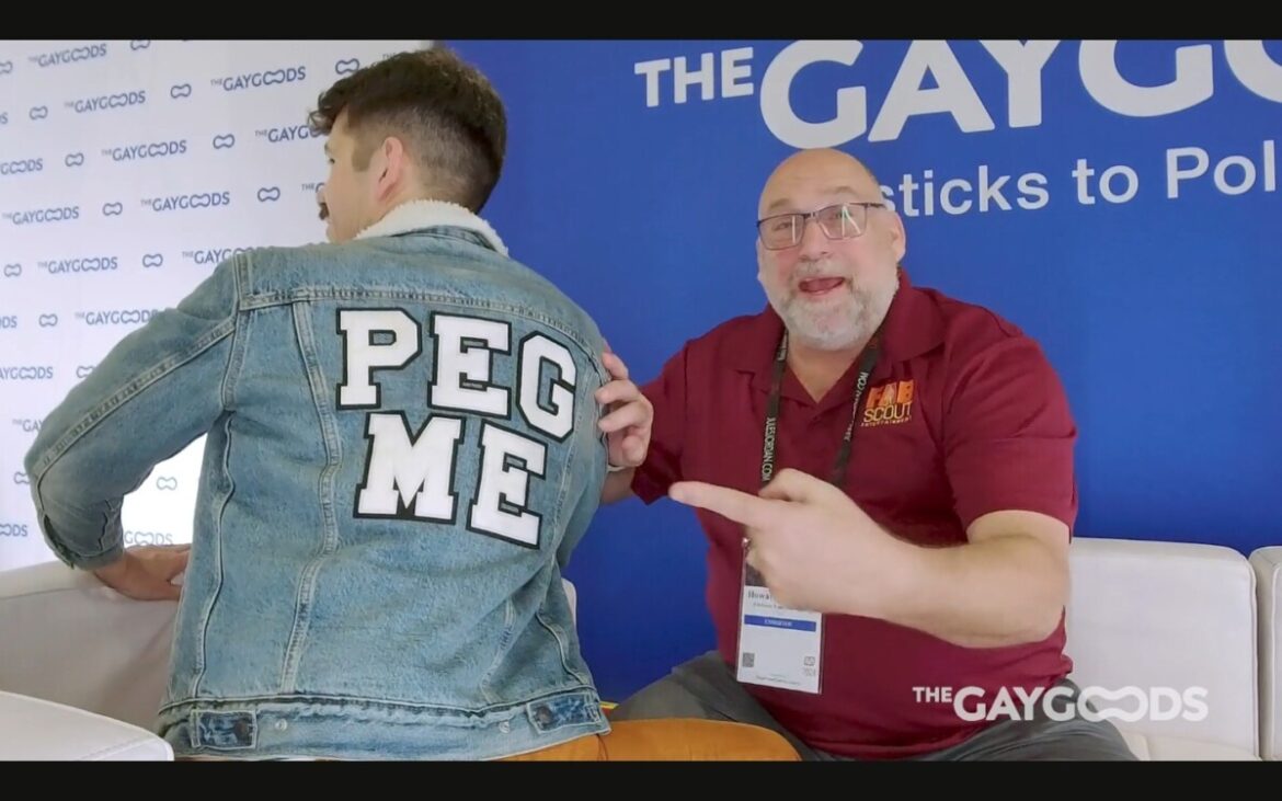 Howard Andrew (a middle aged man with a salt and pepper beard and glasses) is pointing to the back of Jonah Wheeler's jacket. Emblazoned on the back is the words 'PEG ME'. The jacket is denim with a wool lining.