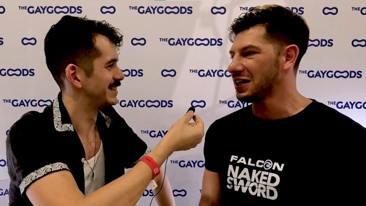 Jonah Wheeler, a caucasian male with a thick moustache is holding a lapel microphone with his right hand and pointing it to Jordan Starr, a beefier man with a black t-shirt saying 'Falcon Naked Sword'
