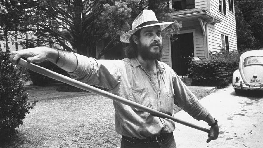 In black and white, a bearded Gene Leggett holding a hockey stick. He wears a fedora, there's a weatherboard house in the background and a Volkswagen Beetle.