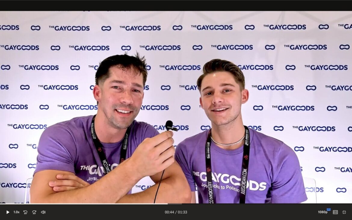 A pair of men, Eddie Patrick (left) and Dex Devall (right) are sitting in front of a white banner saying 'The Gay Goods'. They are both wearing purple T-shirts saying 'The Gay Goods'. Eddie (left) has burly arms, dark hair and heavily lidded eyes. Dex Devall has a bronze complexion is quite young with short golden hair.