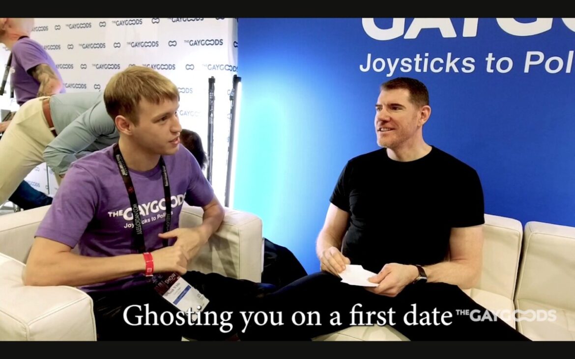 Noah White and Legrand Wolf are sitting on white leather coaches chatting. Noah is a thin young man with blonde hair and a muted gold complexion. He is wearing a purple T-shirt saying "The Gay Goods" in a white font. Legrand Wolf is an older gentleman with dark hair wearing all black (T-shirt and pants).