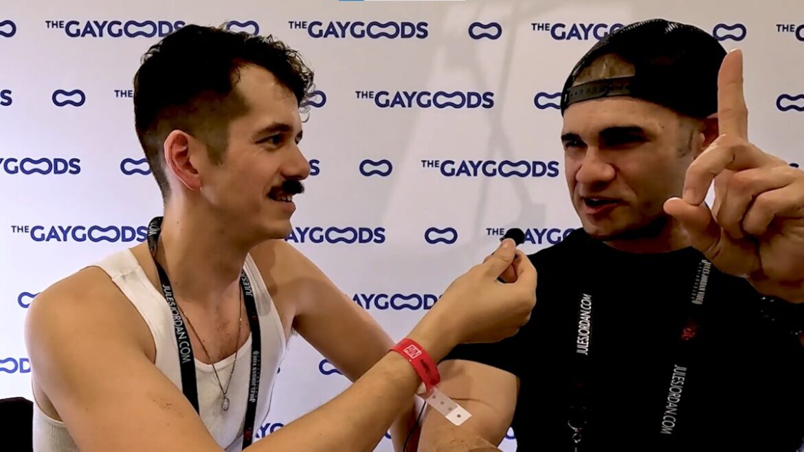 Screenshot 2024 07 30 133851 Jonah Wheeler is speaking with Kyler Drayke at the GayVN awards. Jonah is wearing a white tank top is built lean with dark brown hair and moustache. Kyler Drayke is wearing a black T-shirt and black cap put on backwards. He has a wide nose and a goofy smile. His left hand is pointing up indicating an erection.